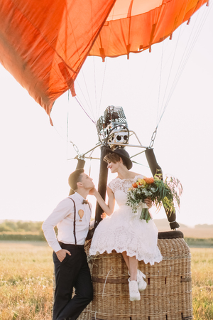 The lovely portrait of the vintage dressed newlyweds spending time in the sunny field. The beautiful bride with the bouquet is sitting on the airballoon basket and petting the face of the groom stending near her.の写真素材