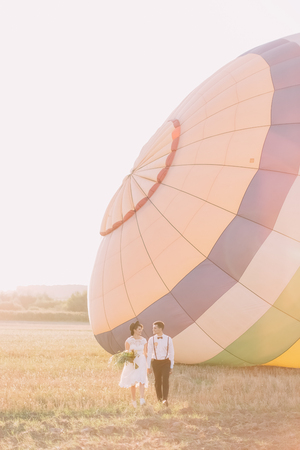 The happy newlyweds holding hands are walking in the sunny field at the background of the colourful airballoon.の写真素材