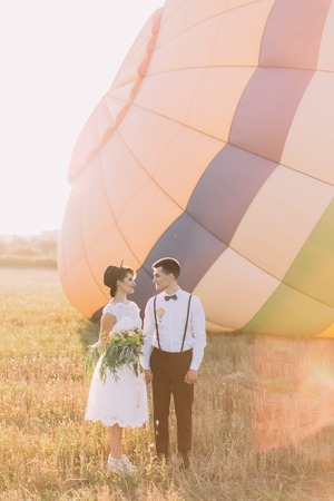 The lovely full-length side portrait of the newlyweds holding hands while the smiling bride is holding the bouquet at the backgrounhd of the airballoon located in the sunny field.の写真素材