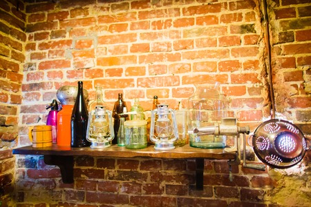 The horizontal view of the wooden shelf with colourful bottle glasses and candle stands hanging near the shining colander on the brick wall pf the cafeの写真素材