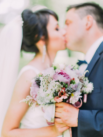 Beautiful wedding bouquet of colourful peonies with herbs in the hands of kissing newlywedsの写真素材