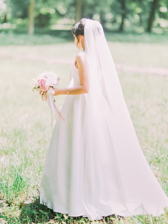 Full-length side view of the bride in the long wedding dress holding the wedding bouquet of peoniesの写真素材