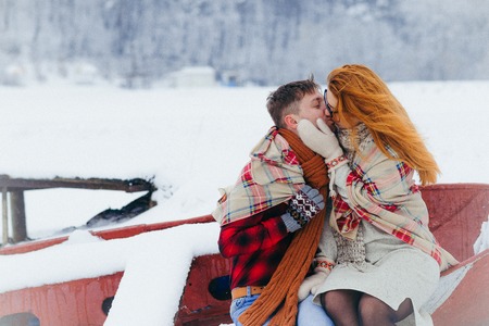 The horizontal portrait of the charming happy couple wrapped in the knitted plaid is kissing while sitting on the boat during the snowfall in the forest.の写真素材