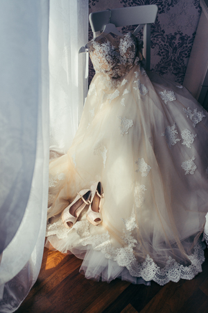 The vertical view of the stylish white long wedding dress with flowers hanging on the chair. The beige high heels are placed on the dress.の写真素材