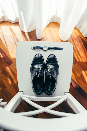 Top view at the stylish black wedding man shoes and hands watch laying on the white chair near the window. Ray of the sun.の写真素材