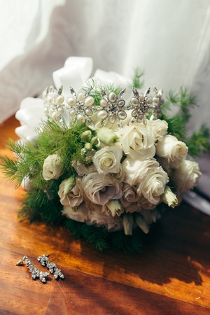 The vertical shot of the long silver earrings with diamonds lying near the wedding bouquet of white roses with the hair hoop placed on it. Isolated composition.の写真素材
