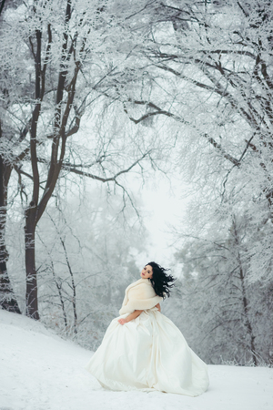 The full-length view of the charming bride with long beautiful black hair standing on the snowy path in the forest.の写真素材