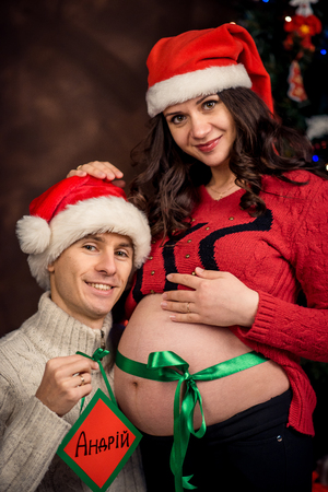 Half-length portrait of the happy couple in Santa Claus hats. The smiling man is hugging his beautiful pregnant wife with wrapped tummy with ribbon and holding the sign Andrii.の写真素材