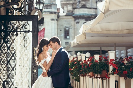 Horizontal wedding portrait of the happy hugging newlyweds in the sunny street. The bride is stroking the cheek of the handsome groom.の写真素材