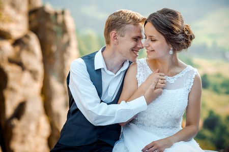 Emotional wedding portrait. Beautiful young newlywed couple is pretty smiling, tenderly holding hands and rubbing noses while sitting on the rocks.の写真素材