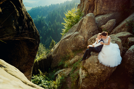 Gorgeous bride is softly hugging the smiling glamour groom back while sitting on the rock at the background of the magnificent landscape of forests.の写真素材
