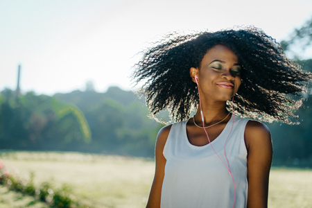 Charming african girl with closed eas and green eye shadows is shaking her curly hair and enjoying the music in her earphones outdoor.の写真素材