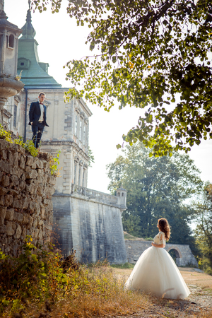 Full-length wedding portrait of the bride looking at the groom standing on the stone wall at the background of the old castle.の写真素材