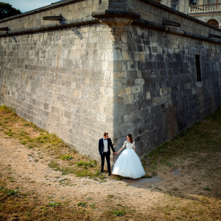 Newlyweds are holding hands while standing in the corner of the huge walls of the old baroque castle.の写真素材