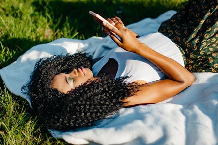 Pretty young african girl is relaxing on the grass. She is texting and browsing via the mobile phone. Above portrait.の写真素材