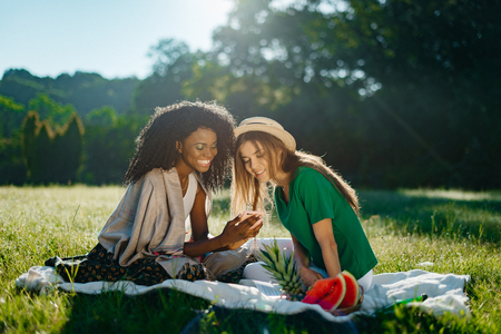 Charming african girl with pretty smile is showing something funny on a phone to her caucasian blonde girlfriend during the picnic on the sunny meadow.の写真素材