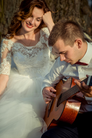The beautiful bride with charming smile is enjoying the guitar play of the handsome groom. Outdoor wedding portrait.の写真素材