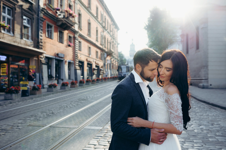 Sunny sensitive wedding portrait. Tenderly hugging beautiful couple of newlyweds in the town street.の写真素材