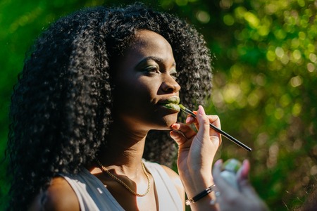 Professional make-up artist is applying the green lipstick on the lips of young charming african girl at the green blurred background.の写真素材