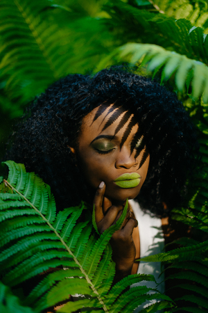 Young alluring african girl with green eyeshadows and lipstick is softly touching her face in the fern garden. Green composition.の写真素材