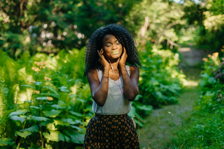 Close-up portrait of the charming peaceful african girl with green lipstick and eyeshadows putting the earphones at the green blurred background.の写真素材