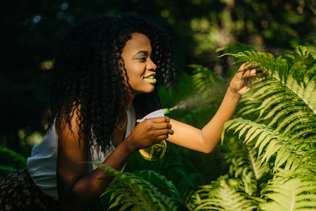 Side outdoor portrait of the attractive young african girl with green lipstick is using the green plastic trigger spray for ferns.の写真素材