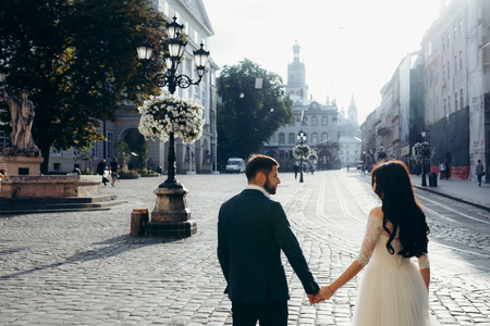 Back half-length view of the gorgeous happy newlywed couple holding hands in the sunny town street.の写真素材