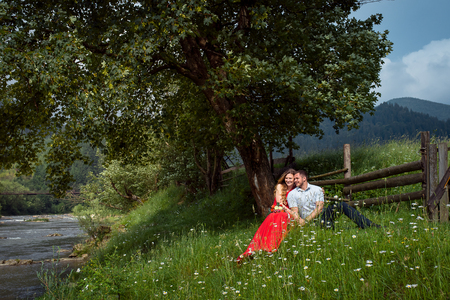The charming smiling couple is hugging while sitting on the green chamomile meadow under the tree near wooden fence and river.の写真素材