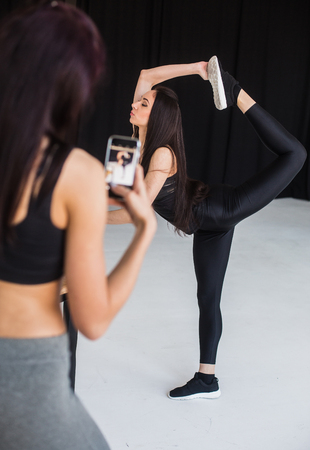 Blurred rear view of the athletic woman taking photo of her sportive girl friend doing the standing bow at the black background.の写真素材
