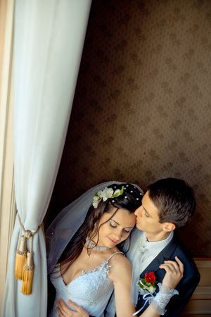 Above romantic wedding portrait. The handsome groom is tenderly hugging and kissing his beautiful brunette bride in the forehead in old rich room.の写真素材