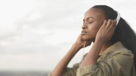 Portrait of the peaceful pretty young african girl with natural make-up enjoying the music in headphones with closed eyes at the blurred white background.の写真素材