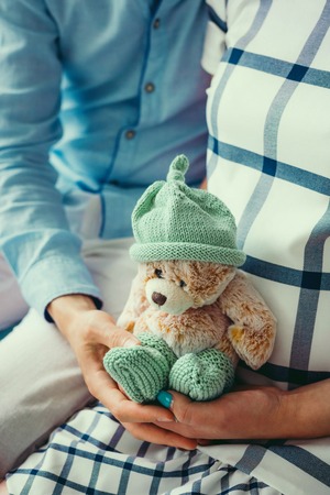 Future parents are holding the small lovely teddy bear in green knitted socks and hat. No face. Close-up shot. Preparation to childbirth.の写真素材