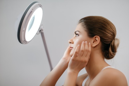 Young fresh woman with blonde hair updo touching her face while looking into cocmetic lamp at white background.の写真素材
