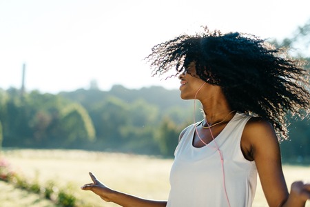 Outdoor emotional portrait of the beautiful young african girl with natural make-up and earphones shaking her dark curly hair on the sunny meadow.の写真素材