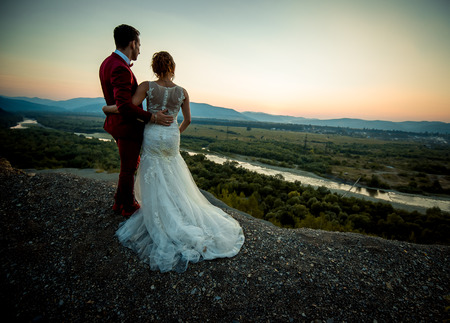 Back view of the beautiful fashionable newlywed couple hugging while standing on the edge of the rock and enjoying the landscape.の写真素材
