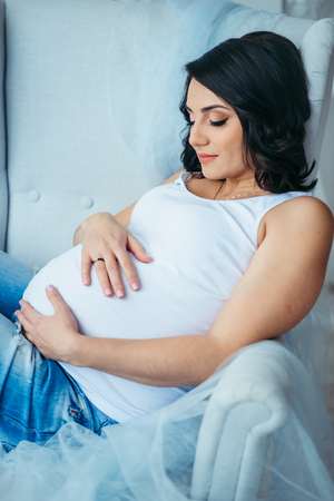 Side portrait of the young attractive pregnant woman touching her stomach while lying in the armchair.の写真素材