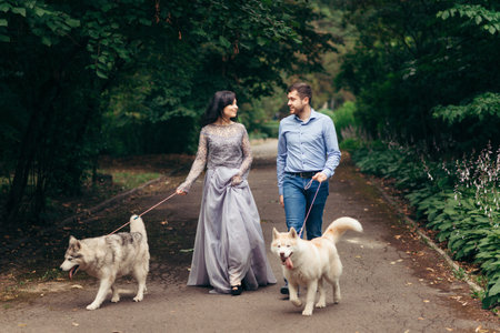 Happy walk of the stylish couple with their two adorable huskies along the green park. The woman is weraing the long evening dress.の写真素材