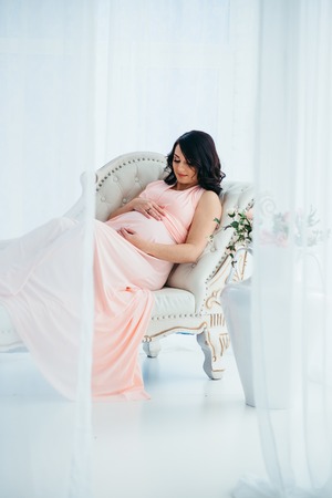 The attractive charming pregnant woman in long pink dress is touching her belly while lying on the sofa near vase with flowers in moder studio.の写真素材