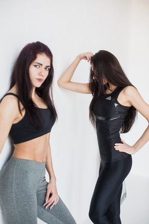 Portrait of two attractive brunette sportwomen in sportwear looking in camera and posing near the white wall. The focus on the woman at the background looking aside.の写真素材