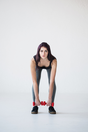 Front full-length view of the beautiful athletic woman bending with the red dumbbells in the studio.の写真素材