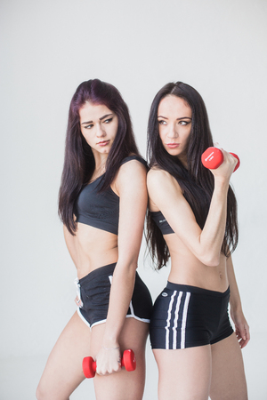 Two young sports women in shot tops and shorts posing with dumbbells and looking aside at white background.の写真素材