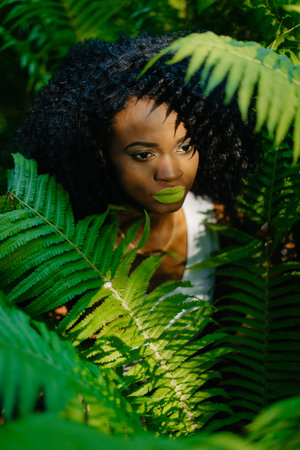 The attractive pretty african girl with green eyeshadows and lipstick is surrounded by green ferns....