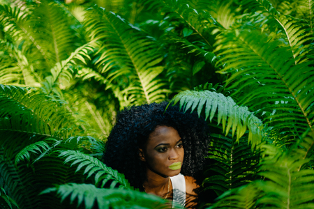 Sensual portrait of the tender gorgeous african girl with green eyeshadows and lipstick looking aside while posing in the fern garden.の写真素材