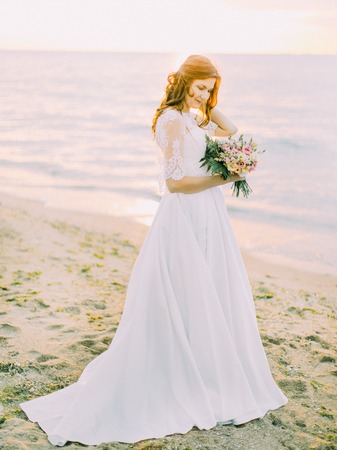 The side view of the bride in the long wedding dress holding the bouquet at the background of the sunset in the sea.の写真素材