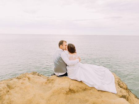 Romantic back portrait of the kissing couple while sitting on the cliff at the background of the sea during the sunset.の写真素材