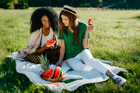 Timespending of two multi-ethnic girl friends.Pretty african girl is chatting via the mobile and eating the watermelon while her attractive blonde friend is reading book and eating fruit.Picnic theme.の写真素材