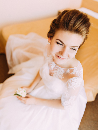 Smiling bride is holding the white mini-bouquet and sitting on the bed. The above portrait.の写真素材