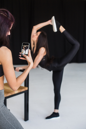 Back view of the brunette taking photo of her attractive athletic girl friend in the pose of standing bow in the studio.の写真素材