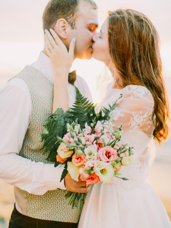 The groom is holding the wedding bouquet of the roses while kissing with the beautiful bride.の写真素材