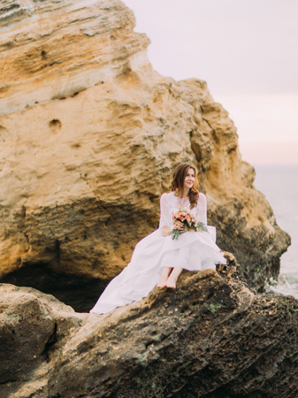 Beautifully dressed bride with the wedding bouquet is sitting on the cliff and looking at the right side at the background of the mountains.の写真素材